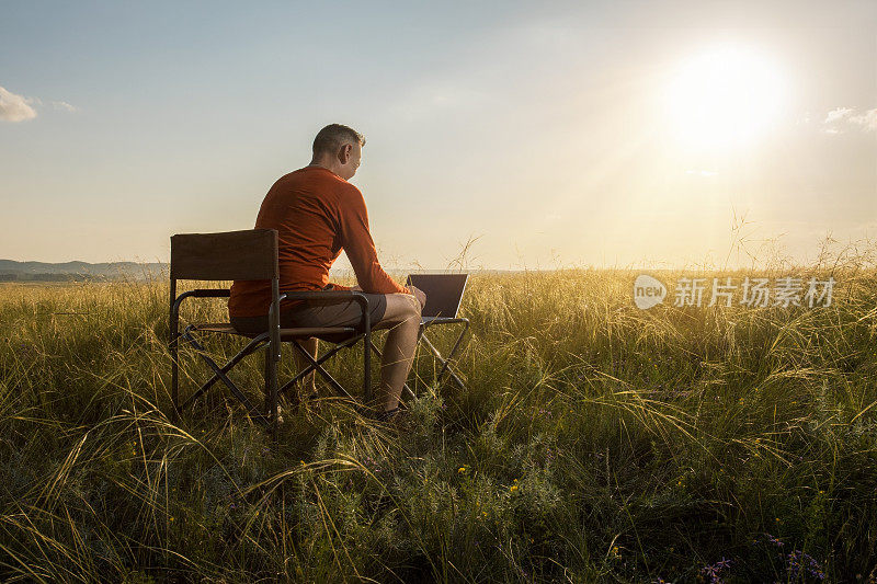 Man traveler博主在上网本电脑上远程工作，同时欣赏日落时户外美丽的自然景观。
